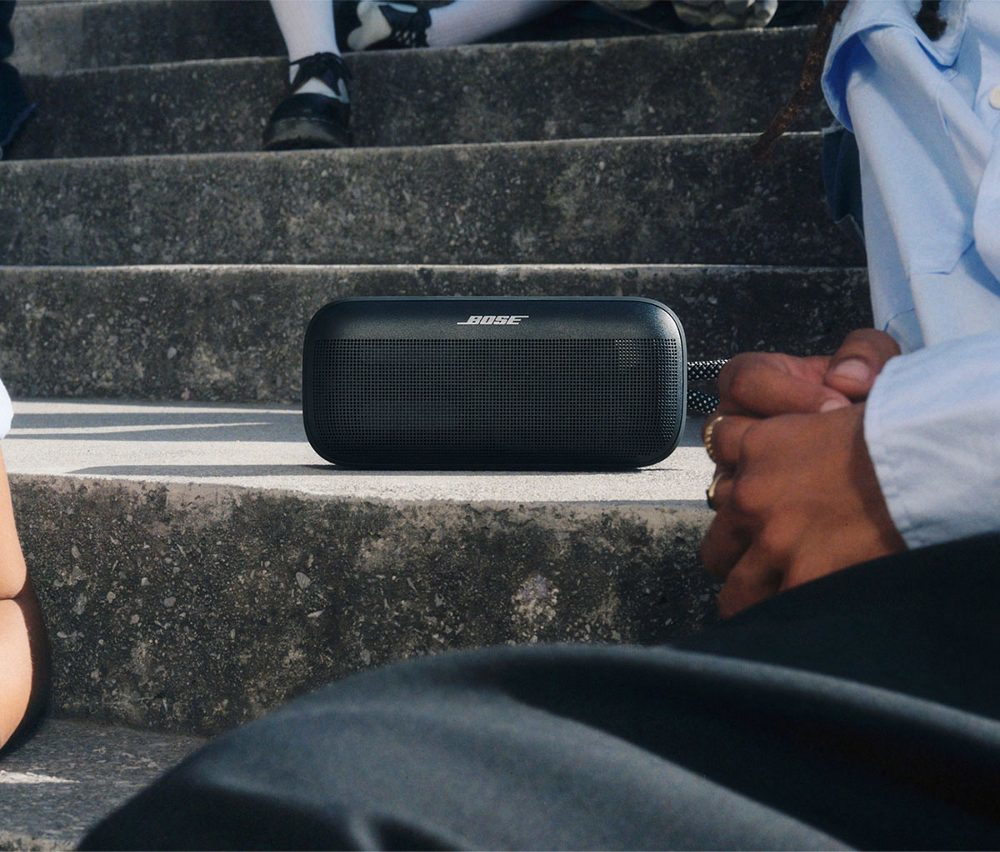Czarny głośnik Bose na betonowych schodach. Ludzie siedzą wokół. Jedna osoba w niebieskiej koszuli. Widoczne nogi i stopy.
