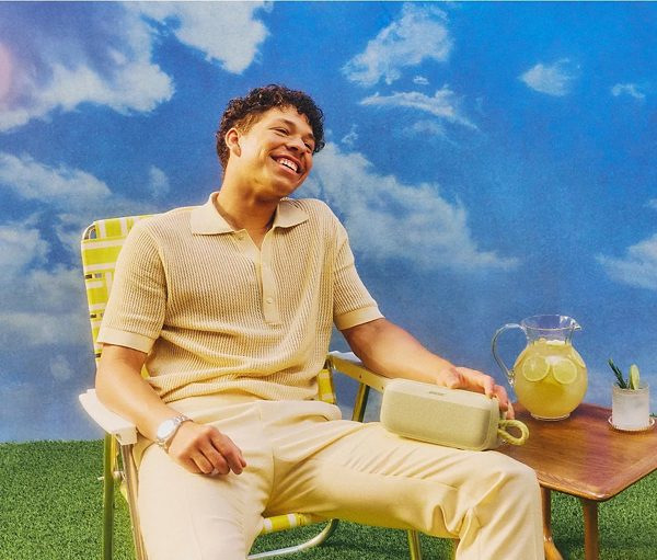 Mężczyzna uśmiecha się siedząc na krześle, trzymając głośnik. Po jego prawej stronie stół z napojem, na tle zachmurzonego błękitnego nieba.