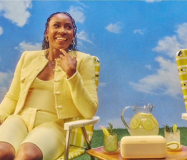 Kobieta w żółtym garniturze uśmiecha się, siedząc na zewnątrz. Napoje i głośnik na stole. Tło nieba z chmurami.