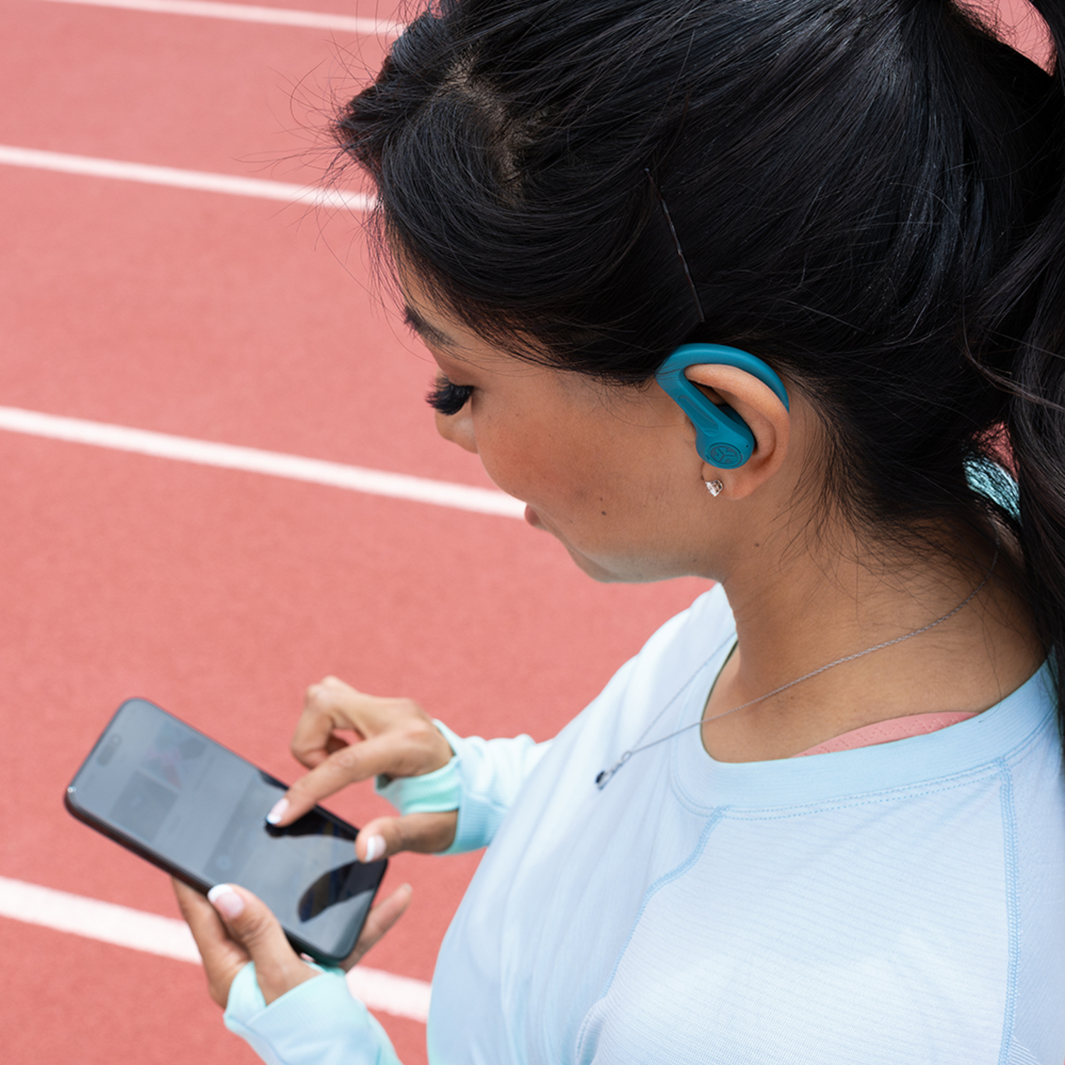 A woman on a track uses her phone. She wears a blue earbud and top. The track is red with white lines.