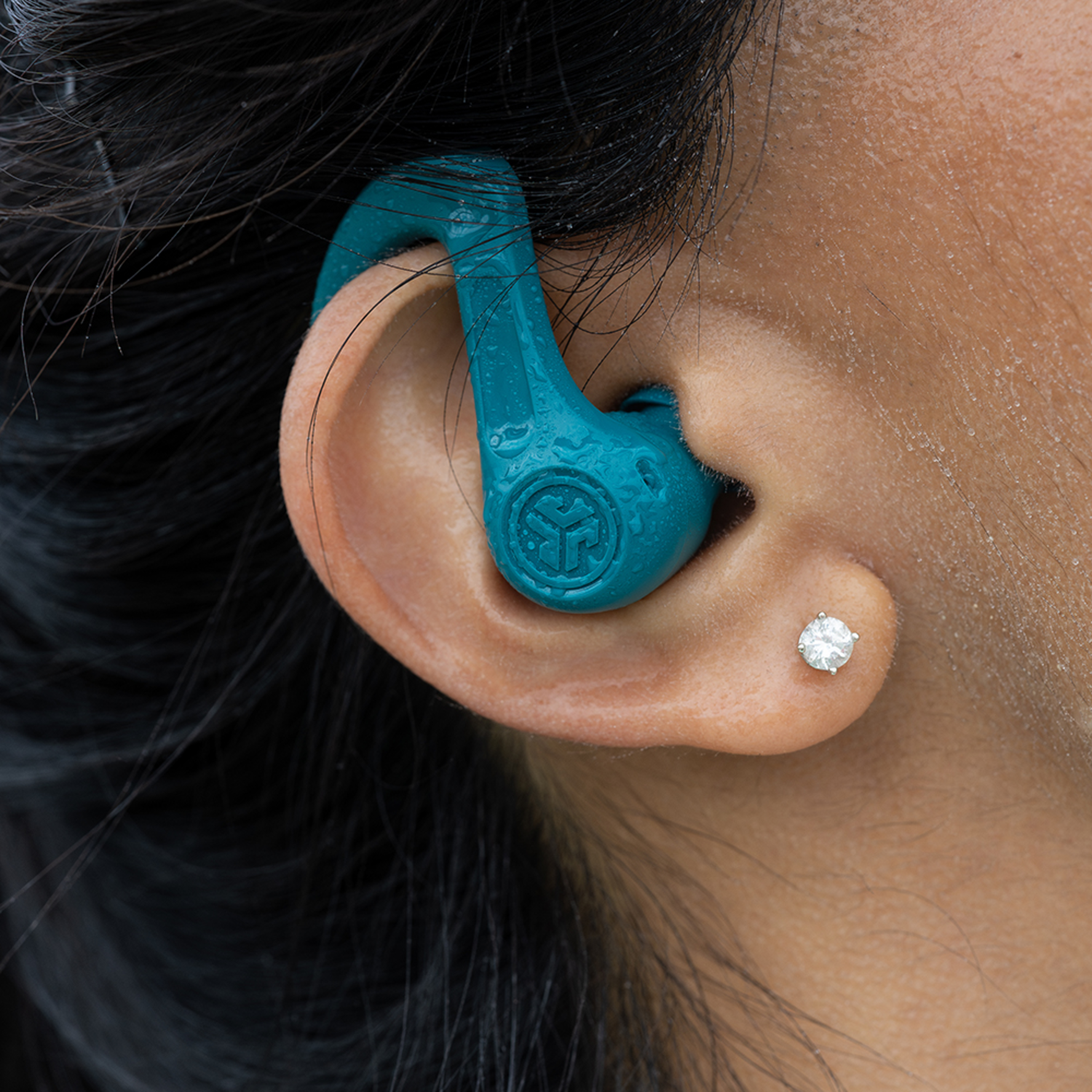 Close-up of ear with teal earbud inserted. Black hair frames the ear. A diamond stud earring is visible.