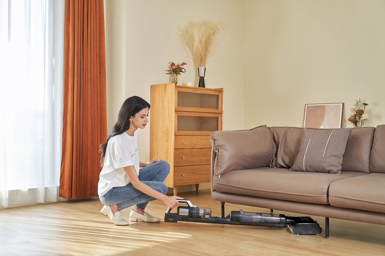 Kobieta klęczy, używając odkurzacza w pobliżu sofy. W tle drewniana szafka i zasłony.