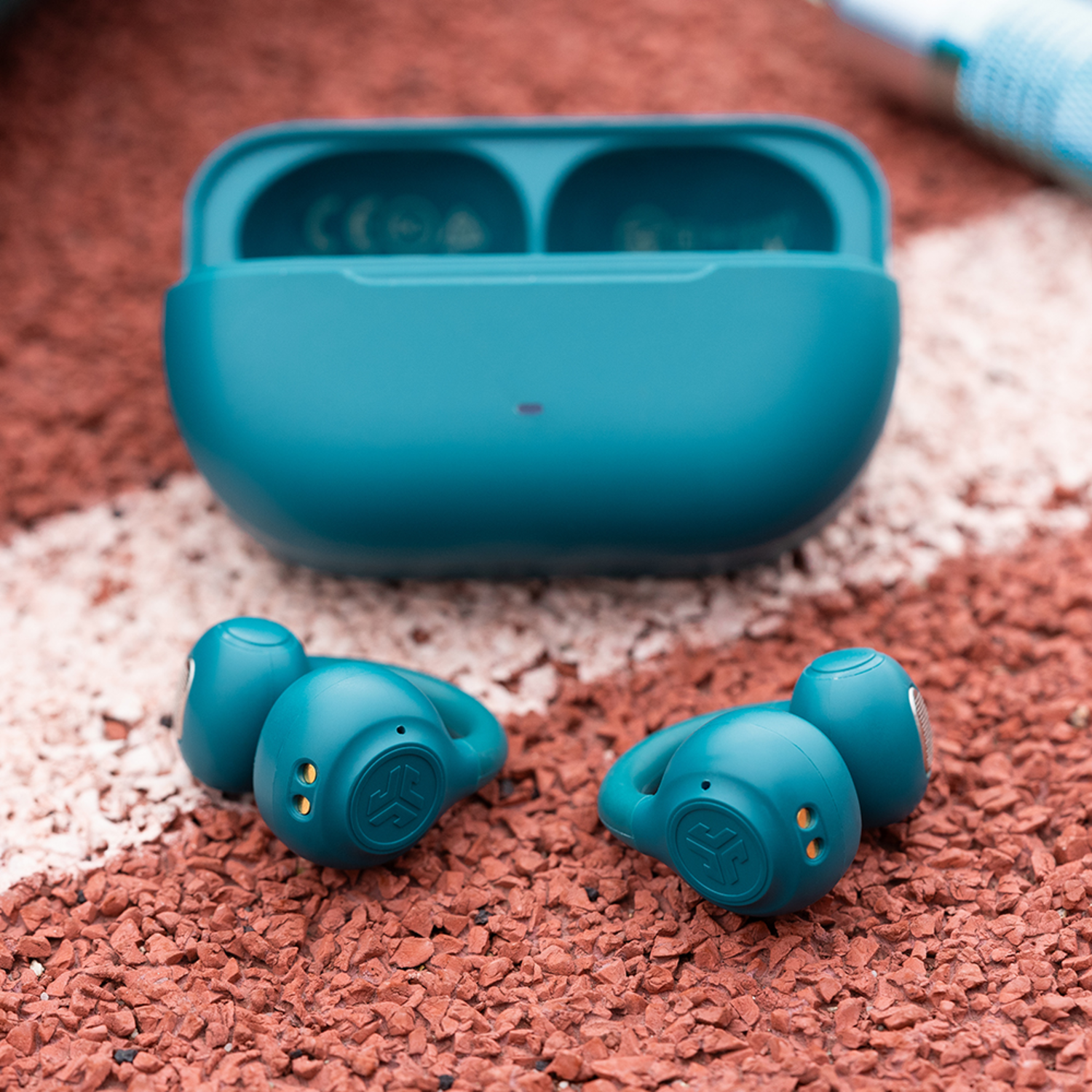 Teal earbuds and charging case on a red speckled surface with a white stripe. A white and blue cloth is in the background.
