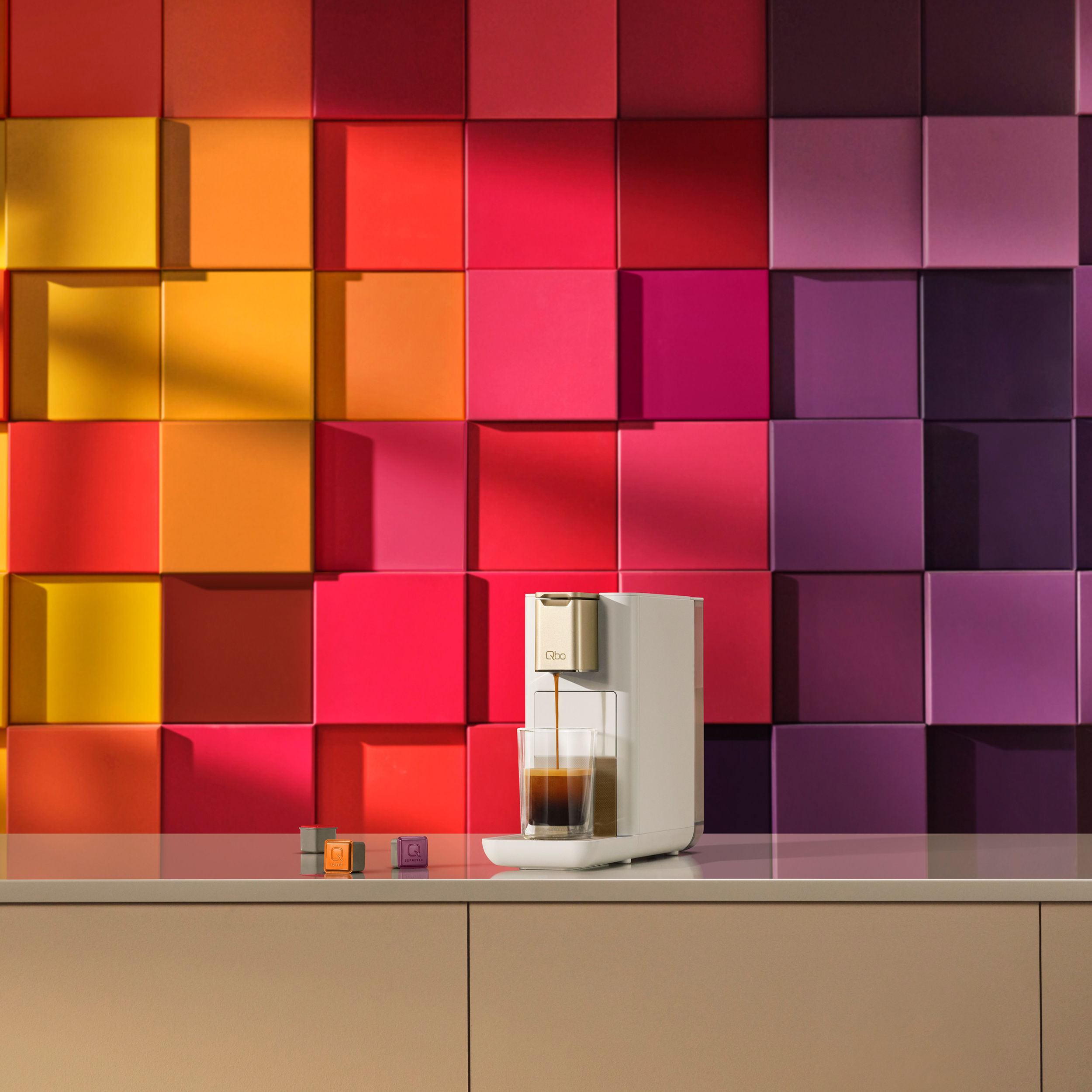 Eine weiße Kaffeemaschine gibt Espresso in ein Glas aus. Der Hintergrund hat bunte quadratische Fliesen. Drei Kapseln auf der Theke.
