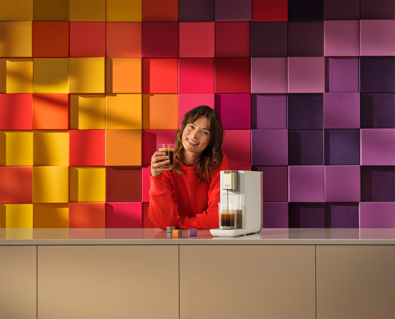 Frau im roten Pullover hält Kaffee. Kaffeemaschine rechts. Bunter Würfelhintergrund. Beiger Tresen.