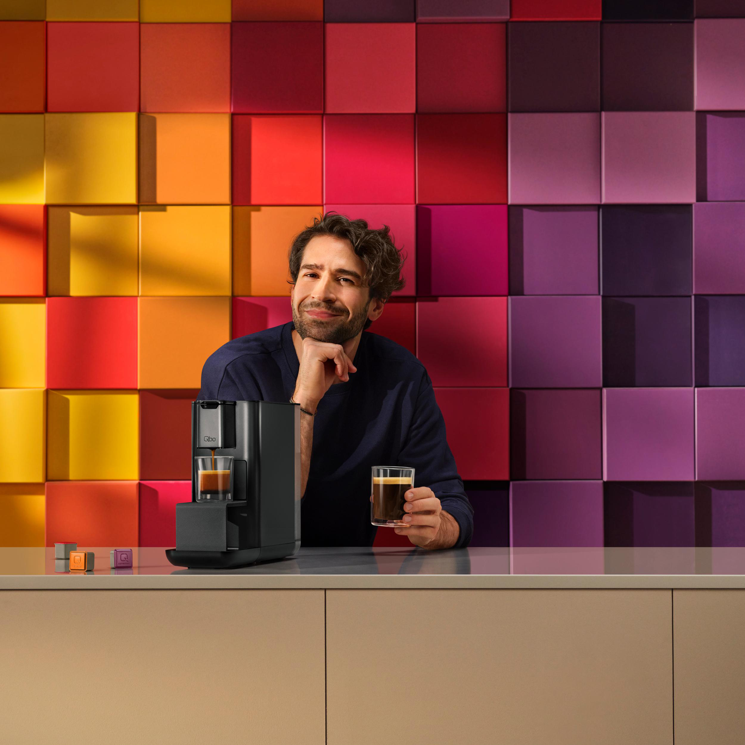 Ein Mann mit dunklen Haaren hält Kaffee. Hinter ihm eine bunte Wand und eine Kaffeemaschine.