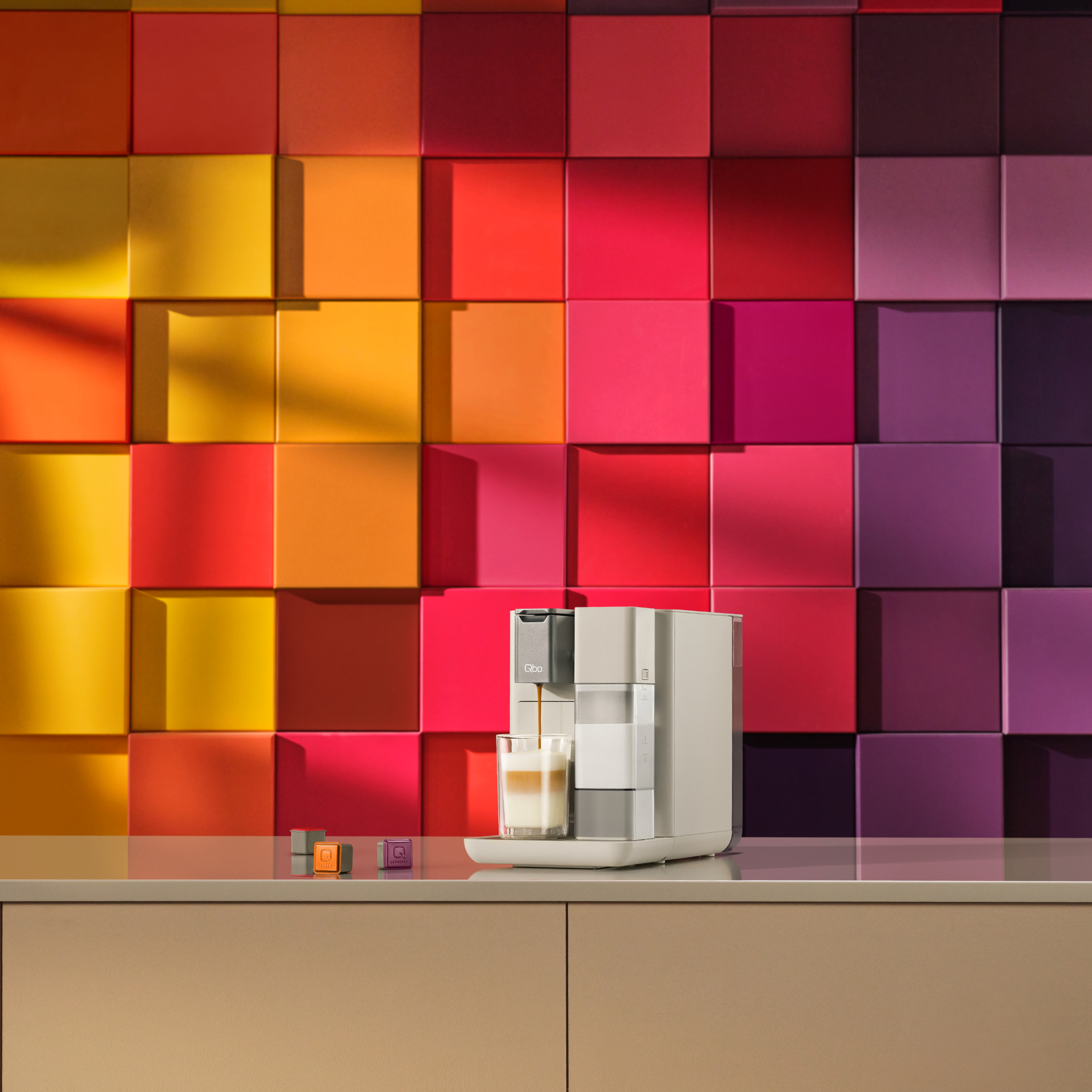Eine Kaffeemaschine, die Kaffee in ein Glas ausgibt. Bunte quadratische Wand im Hintergrund. Kapseln auf der Theke.