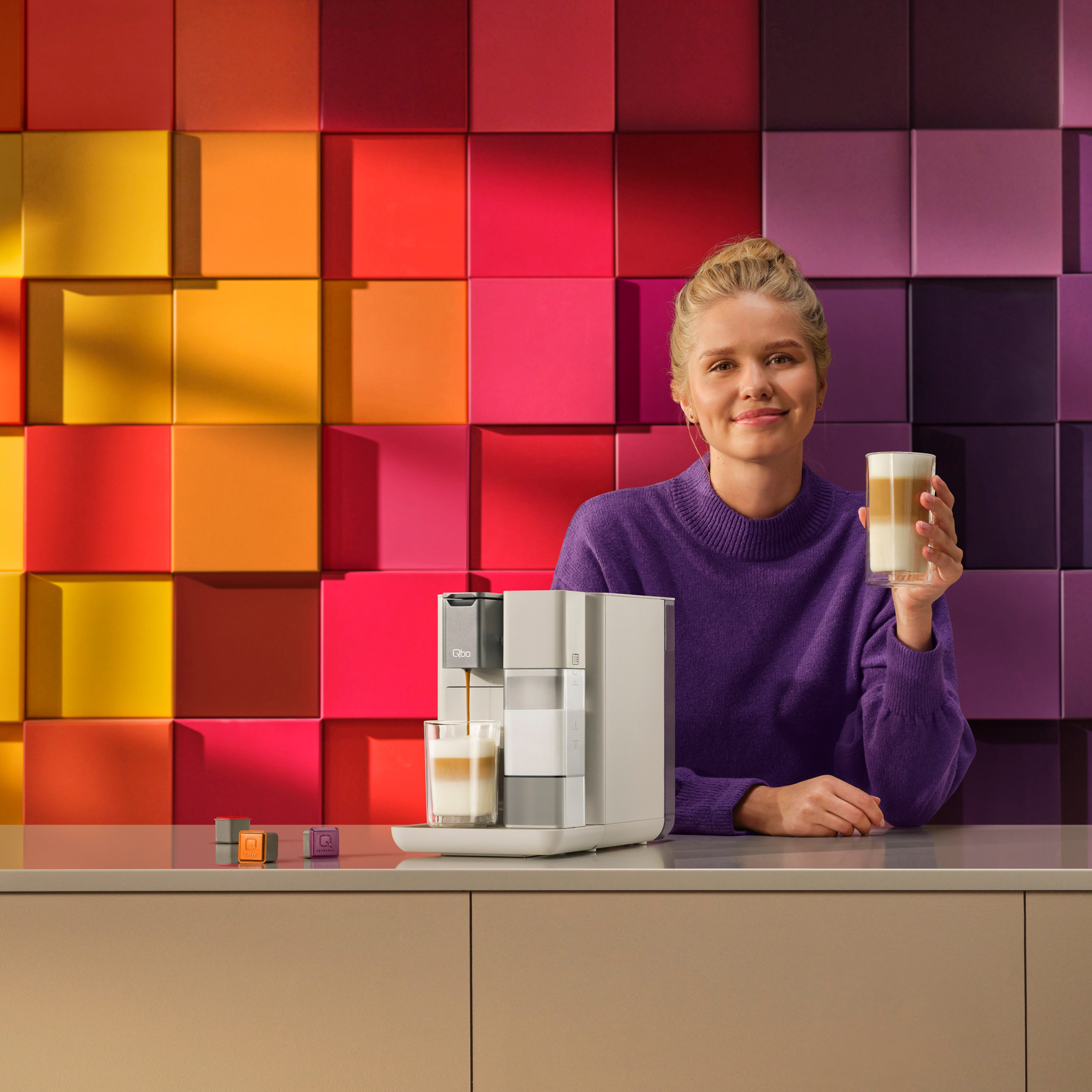 Eine Frau hält einen Latte und lächelt. Eine Kaffeemaschine gießt. Bunte quadratische Wand im Hintergrund. Kaffeekapseln auf der Theke.