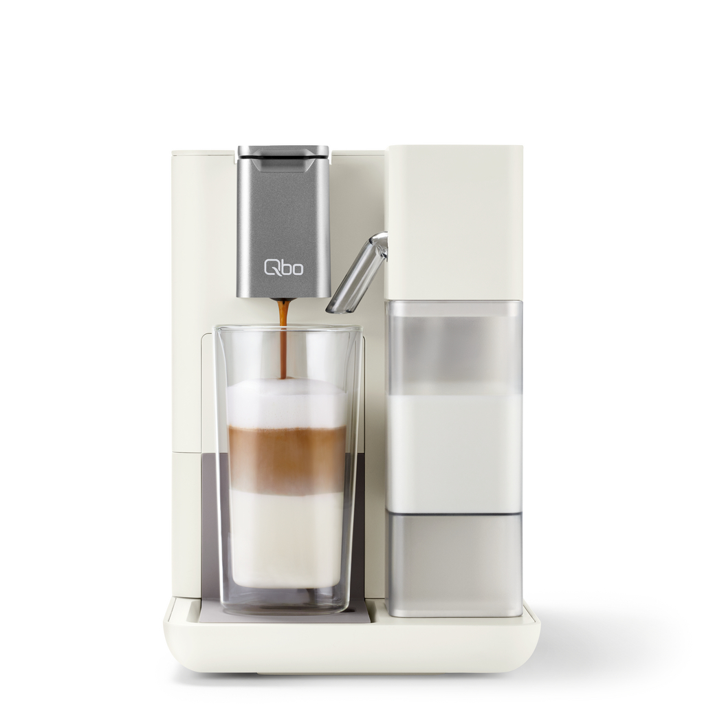 Eine Kaffeemaschine, die Kaffee in ein Glas mit Milch und Schaum schüttet. Die Maschine ist weiß und grau.
