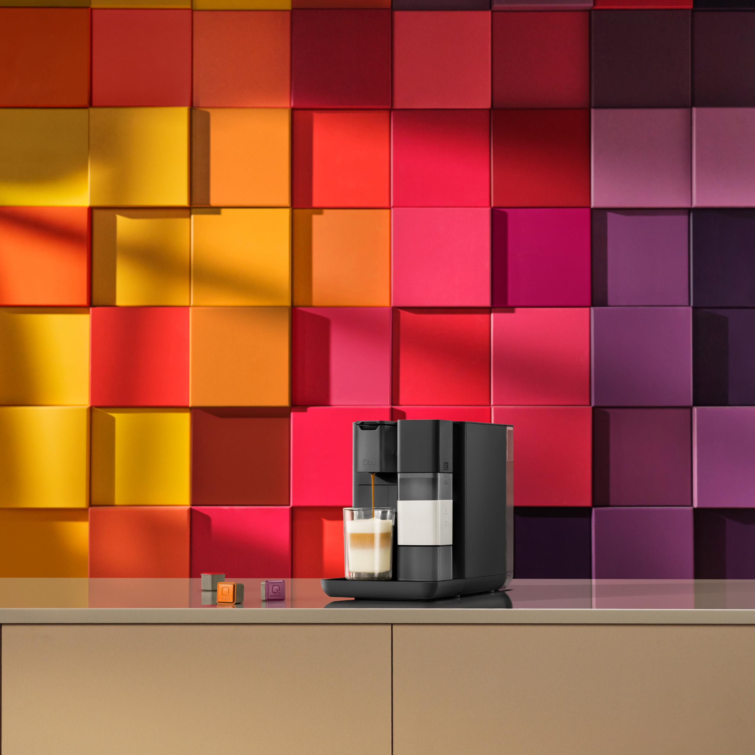 Eine schwarze Kaffeemaschine gießt Kaffee in ein Glas. Bunte quadratische Wand im Hintergrund. Drei Kaffeekapseln davor.