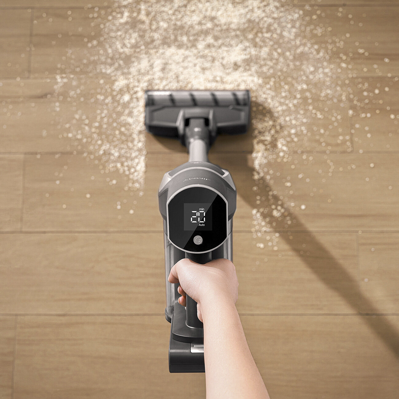 Dłoń trzyma odkurzacz czyszczący drewnianą podłogę. Pył jest rozrzucony wokół odkurzacza. Wyświetlacz pokazuje 20 min i Auto.