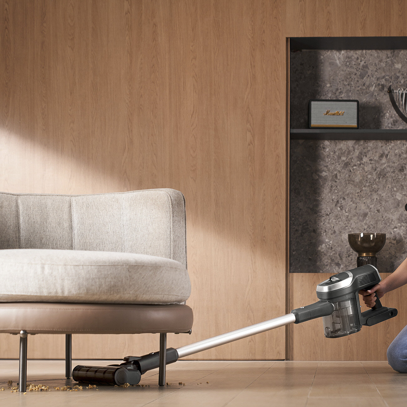 Osoba odkurza drewnianą podłogę. Używany jest szary odkurzacz. Po lewej stronie znajduje się jasno kolorowa kanapa. W tle głośnik.