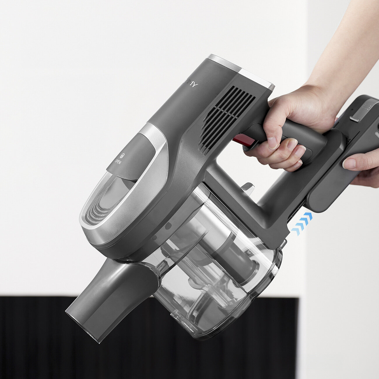Ręka trzyma szary odkurzacz bezprzewodowy. Pojemnik na kurz jest przezroczysty. Tło jest białe i czarne.