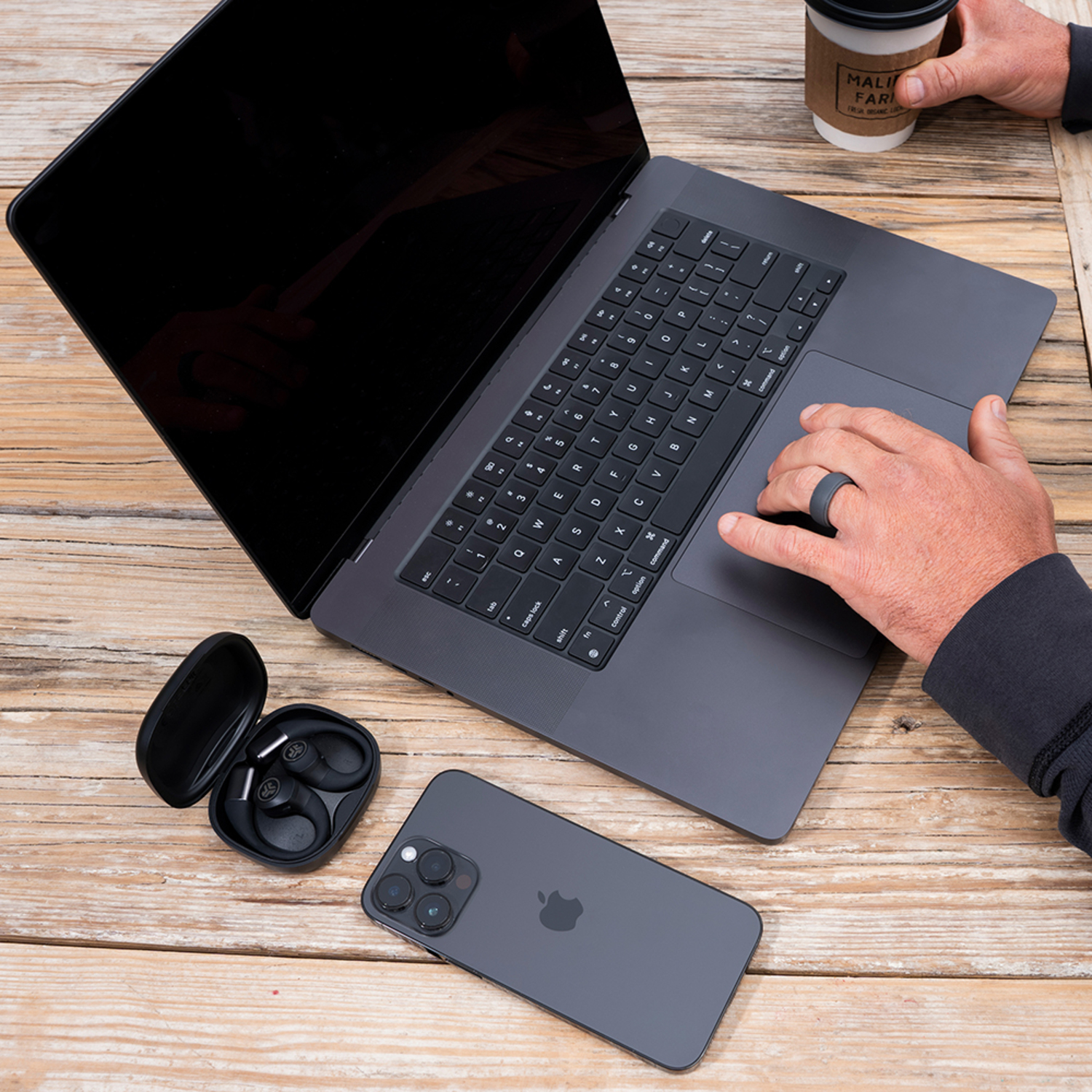Un ordinateur portable, un téléphone et des écouteurs reposent sur une table en bois. Les mains d'une personne utilisent l'ordinateur portable et tiennent une tasse de café.