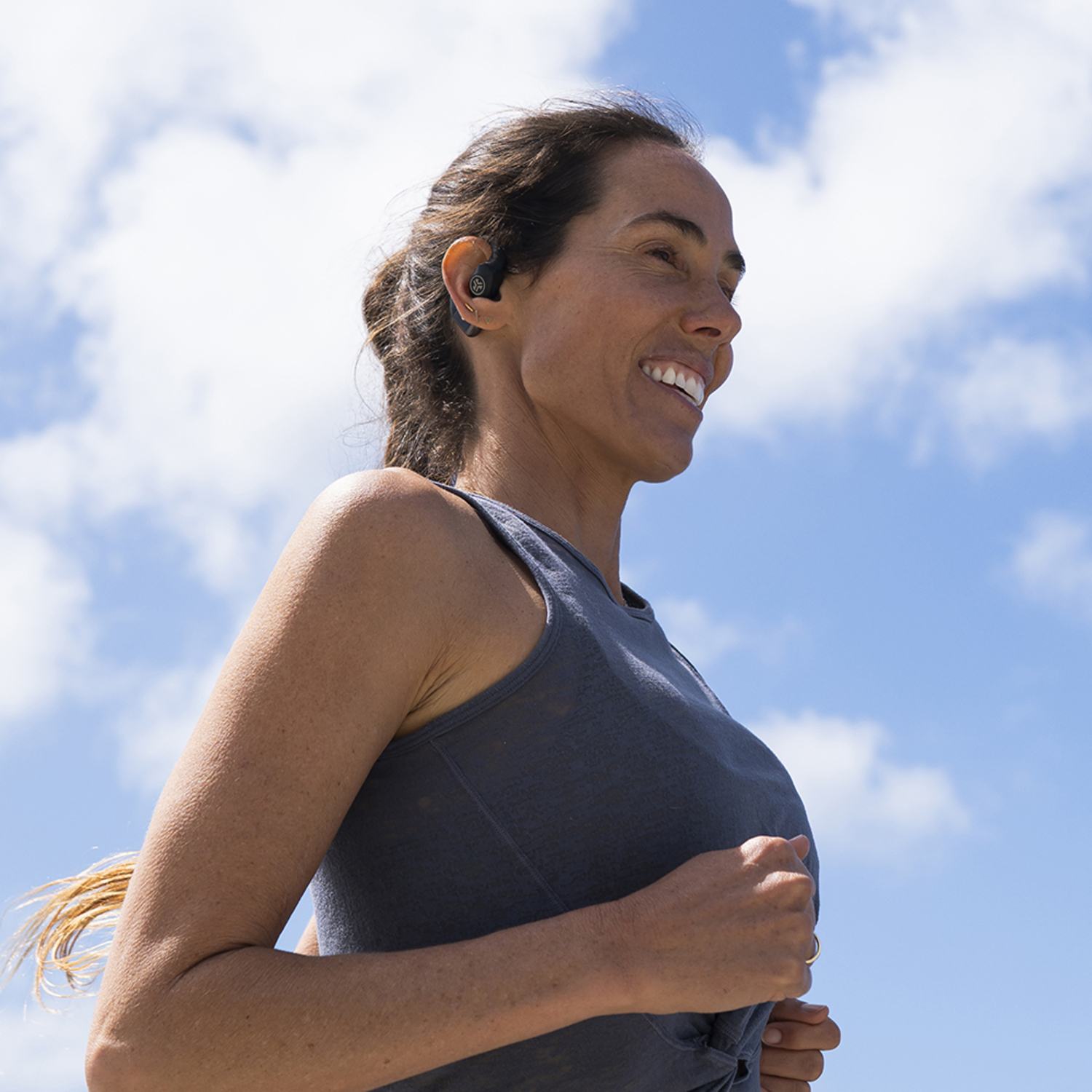 Une femme en débardeur gris court dehors en souriant. Elle porte des écouteurs. Le ciel est bleu avec des nuages blancs.