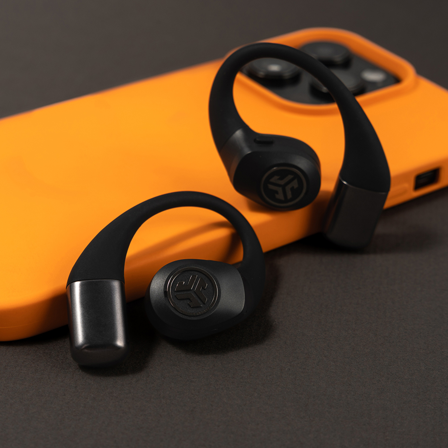 Des écouteurs sans fil noirs avec des crochets d'oreille reposent sur une coque de téléphone orange sur fond gris foncé.