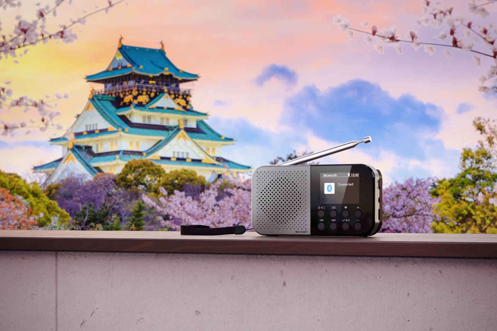 Radio stoi na drewnianej powierzchni z zamkiem Osaka w tle. Obecne są kwiaty wiśni i błękitne niebo z chmurami.