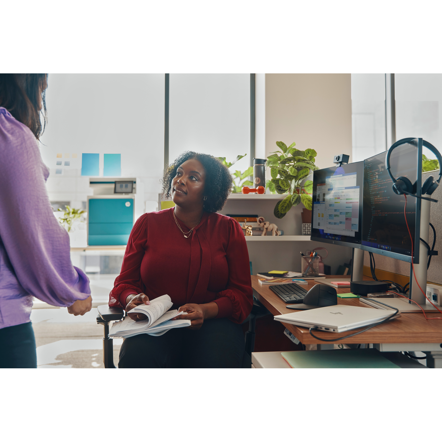 Twee vrouwen in een kantoor. Eén in het rood houdt documenten vast, de ander in het paars. Computermonitoren, planten en een laptop op het bureau.
