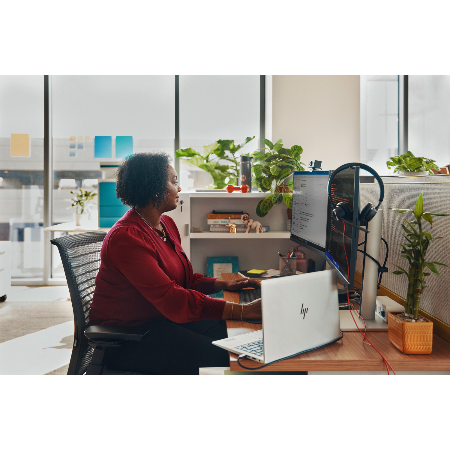 Een vrouw in een rood overhemd zit aan een bureau en werkt op een laptop en twee monitoren. Planten, koptelefoon en een raam zijn zichtbaar.