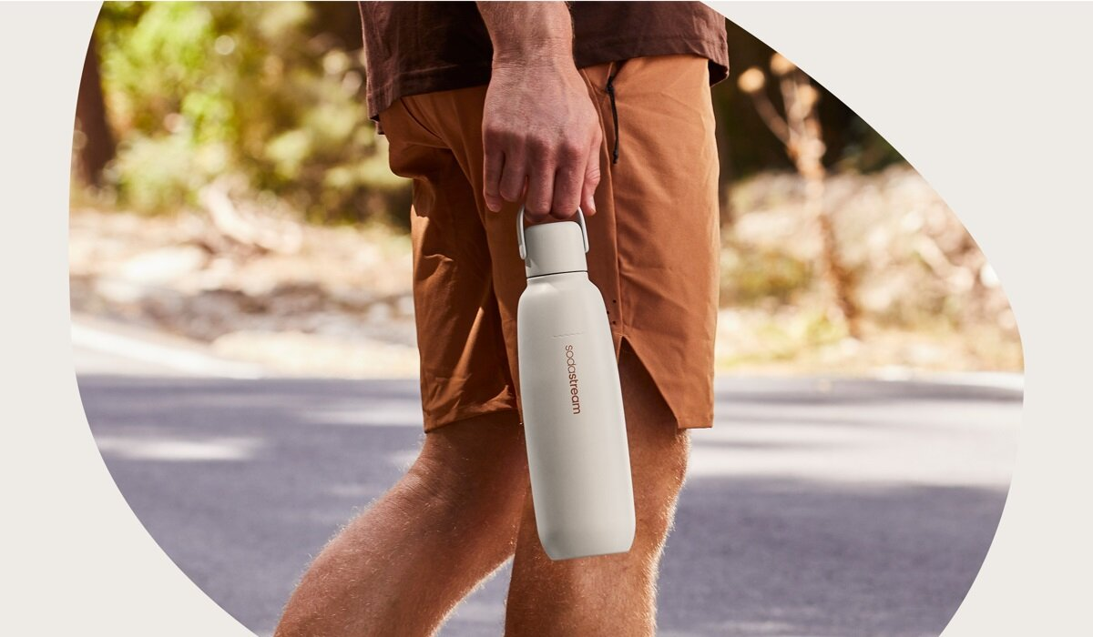 Osoba w szortach trzyma białą butelkę na wodę SodaStream. Tło przedstawia drogę i rozmyte zielone liście.