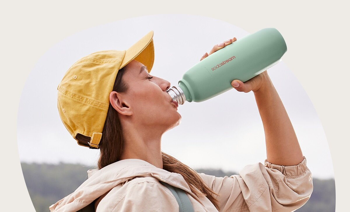 Kobieta pije z zielonej butelki SodaStream. Ma na sobie żółtą czapkę i beżową kurtkę. Tło jest rozmyte.