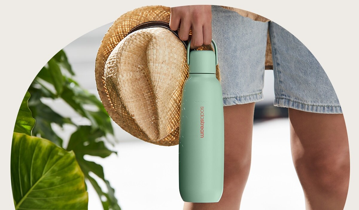 Osoba trzyma słomkowy kapelusz i zieloną butelkę z wodą. Widoczne są dżinsowe szorty. Bujne zielone liście w tle.