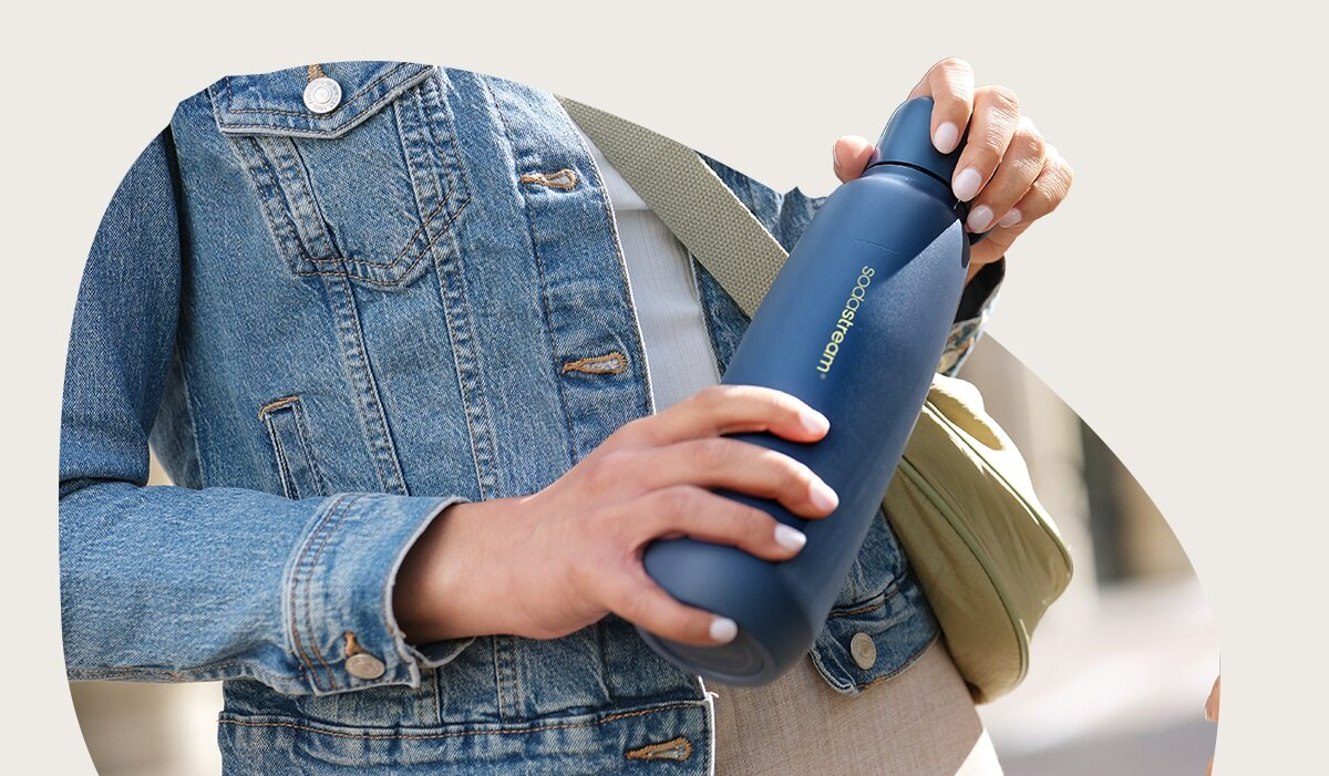 Osoba trzyma niebieską butelkę z wodą. Ma na sobie dżinsową kurtkę i białą koszulę. Zielona torba jest na ramieniu.