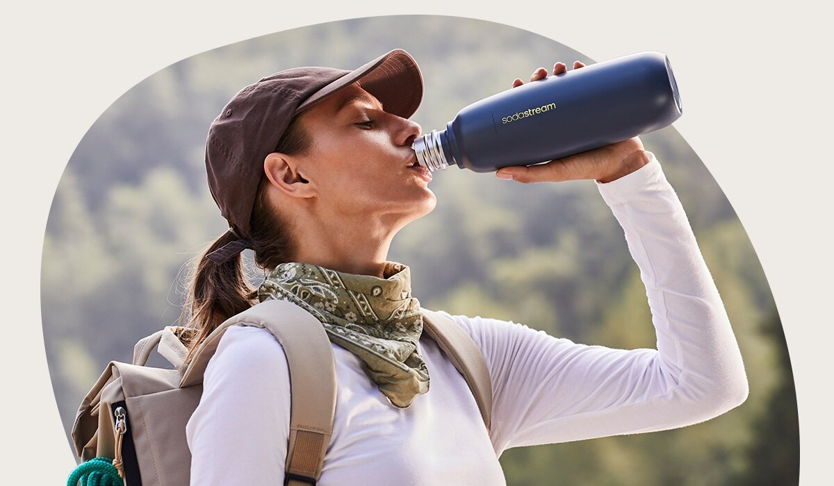 Kobieta pije wodę z ciemnoniebieskiej butelki. Ma na sobie brązową czapkę, bandanę i plecak, z rozmytym tłem.