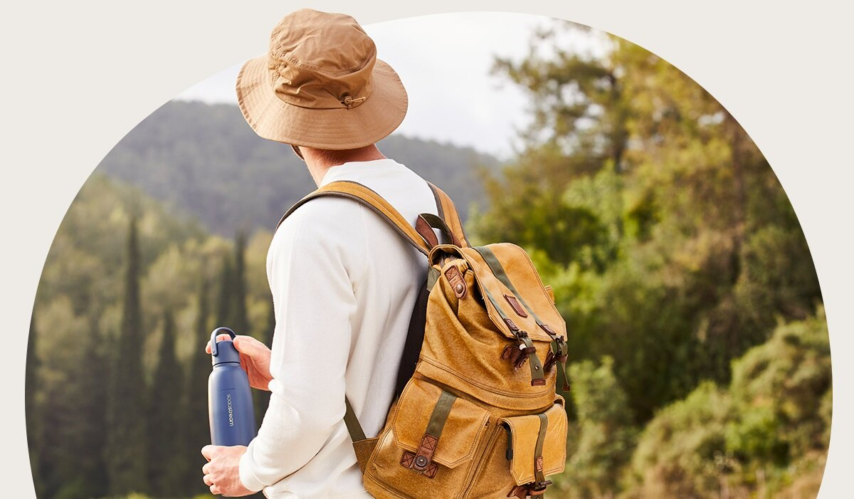 Osoba w kapeluszu i plecaku stoi na łonie natury trzymając butelkę z wodą, tyłem do widza.