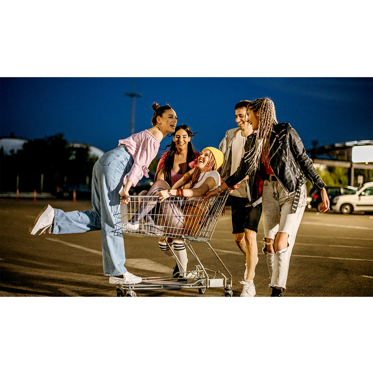Grupo de amigos riendo, empujando un carrito de compras con una persona dentro, en un estacionamiento de noche.