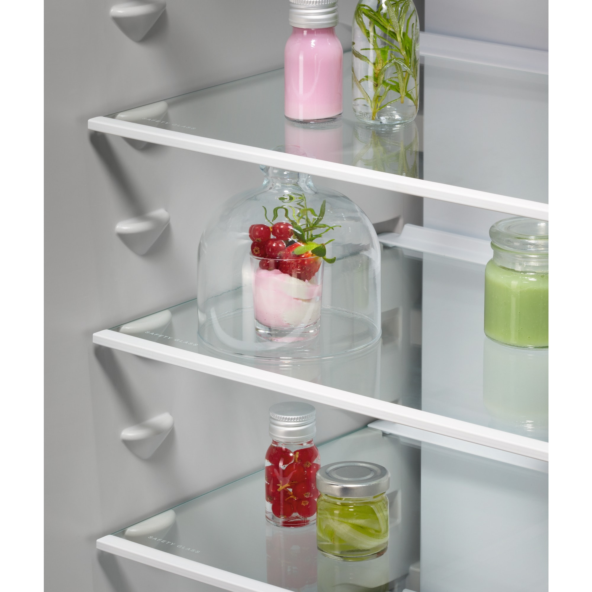 Kühlschrankinnenraum. Glasböden mit Gläsern mit rosa Flüssigkeit, grünen Gegenständen und roten Beeren. Eine Glaskuppel bedeckt ein Dessert.