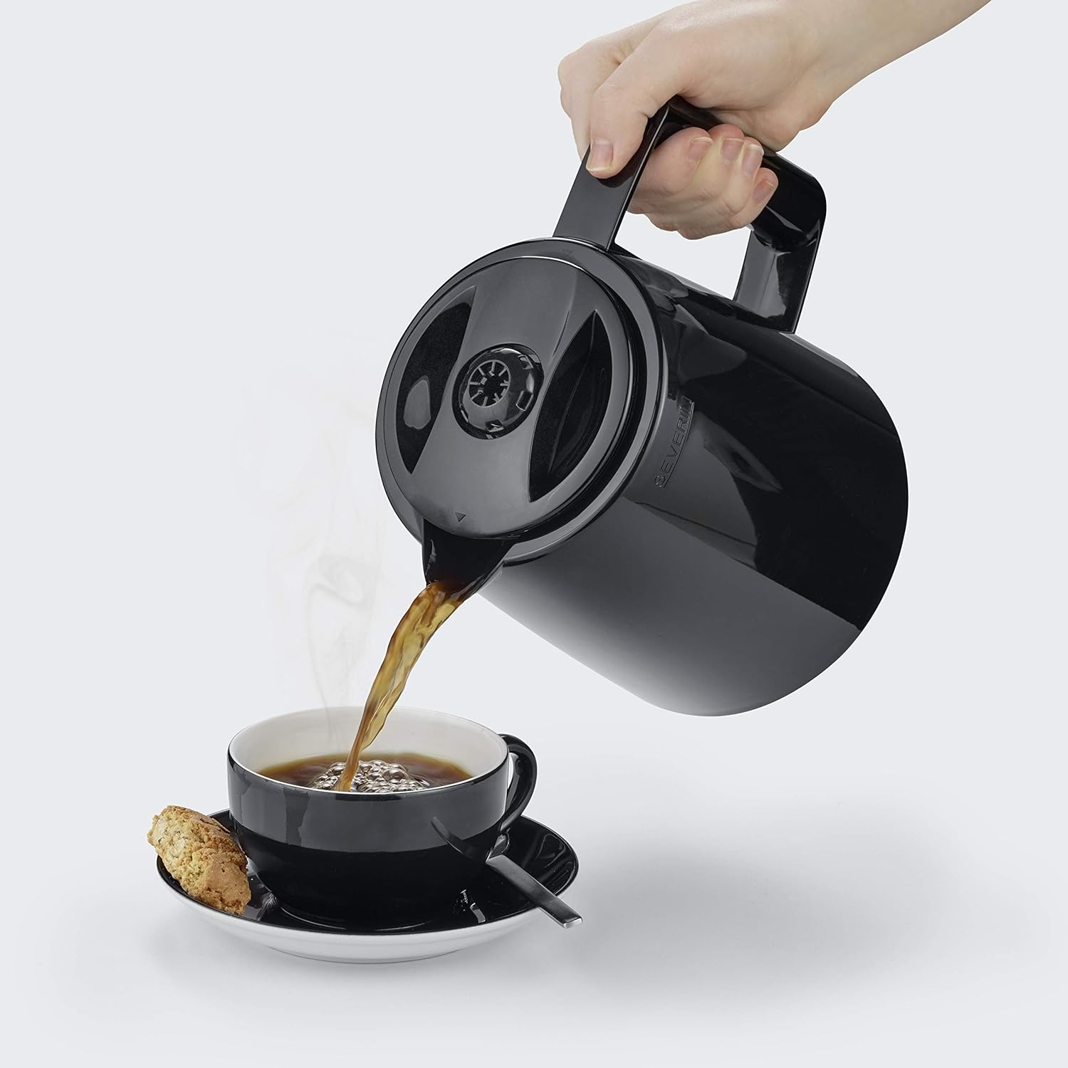 Ręka nalewa kawę z czarnej karafki do czarnego kubka na spodku. Obok kubka leży ciastko. Para unosi się w górę.