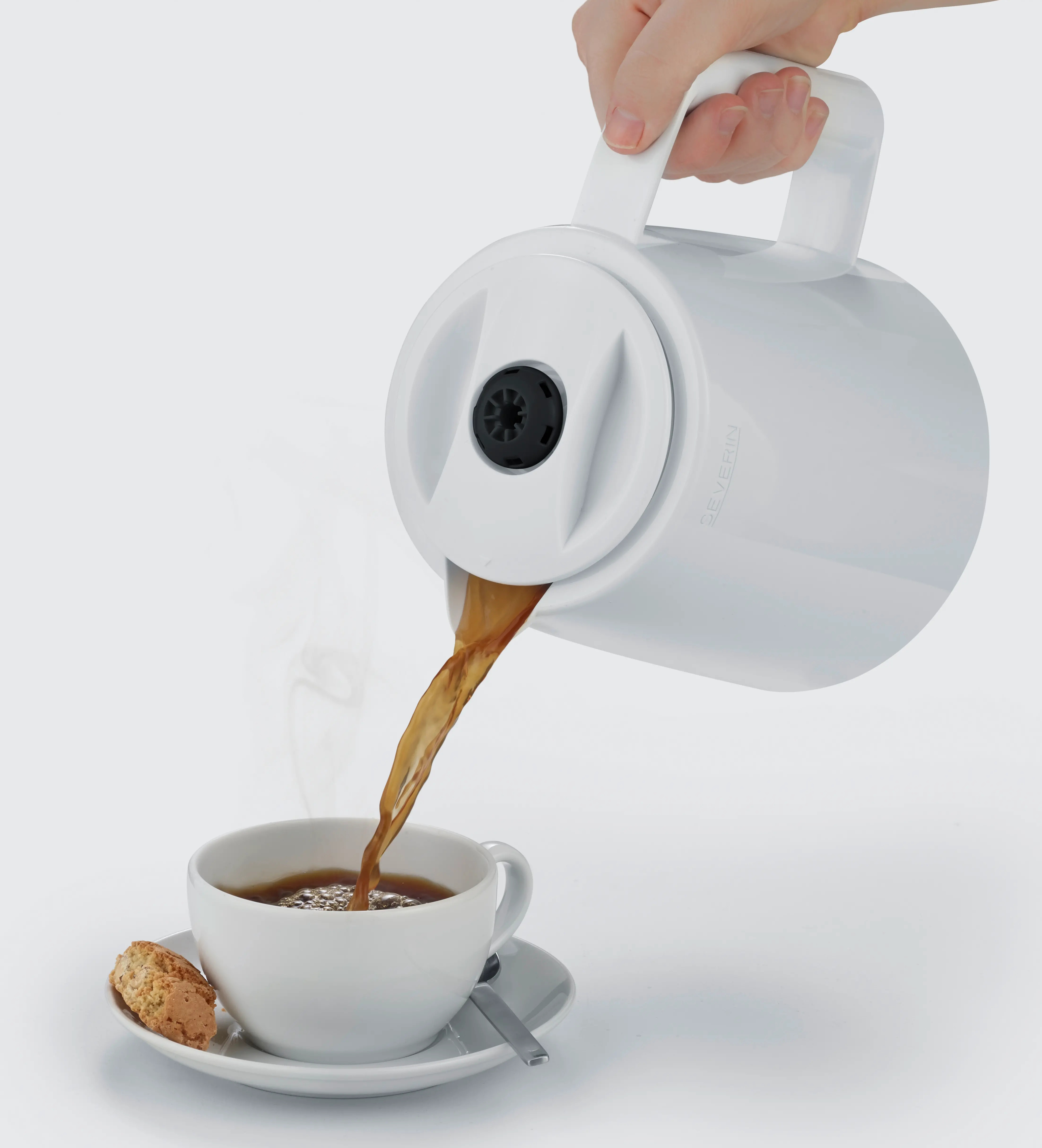 Biały dzbanek do kawy wlewa kawę do białego kubka na spodku z ciastkiem. Ręka trzyma dzbanek.