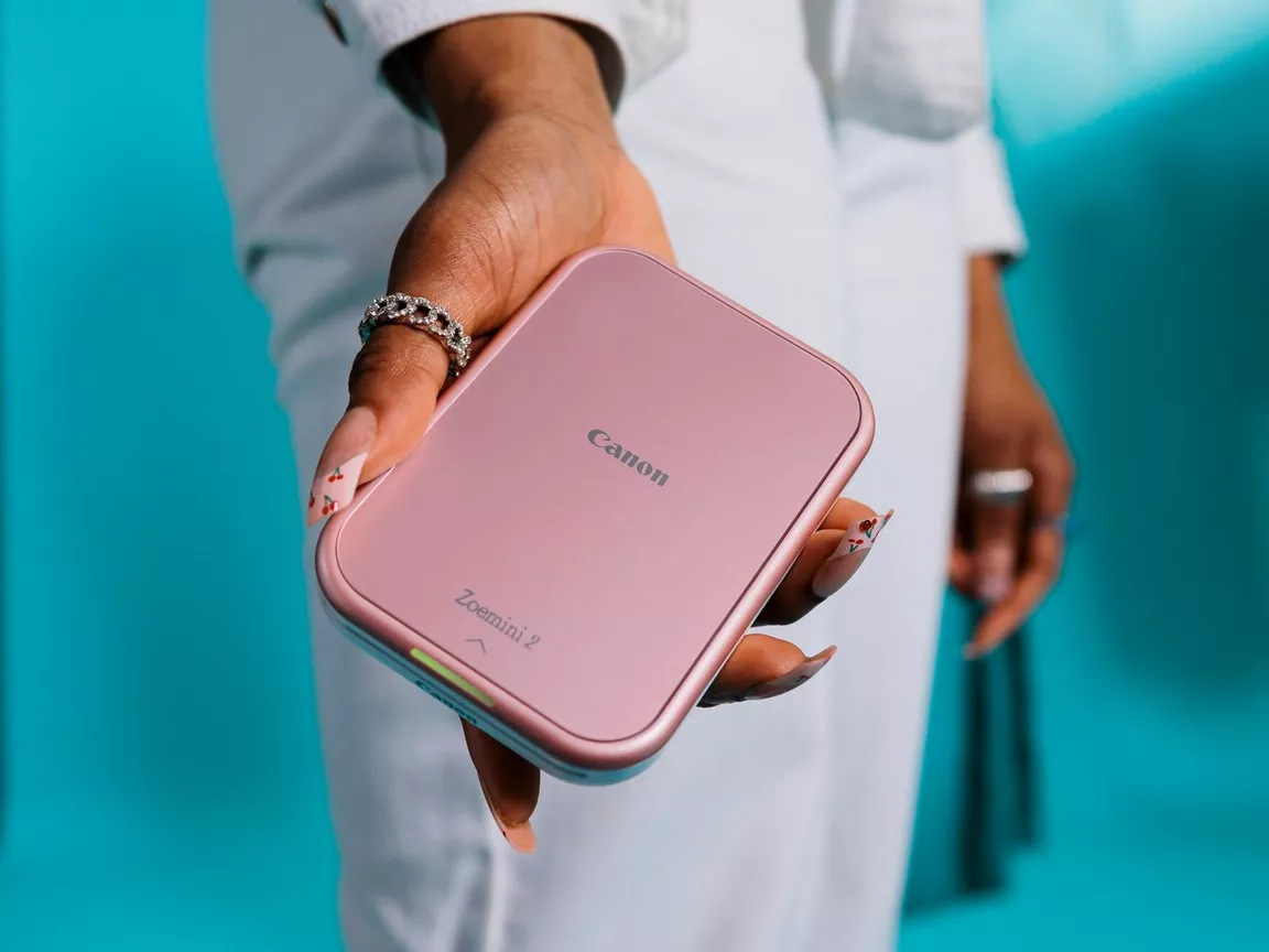 Osoba trzyma różowy Canon Zoemini 2. Osoba nosi pierścionek, ma długie paznokcie i białą koszulę. Niebieskie tło.