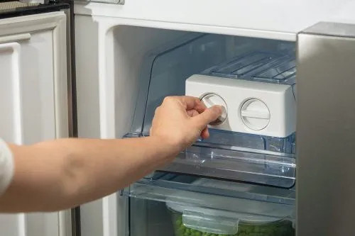 Ręka reguluje pokrętło wewnątrz lodówki. Widoczny biały moduł sterujący. Zielony groszek w pojemniku poniżej.