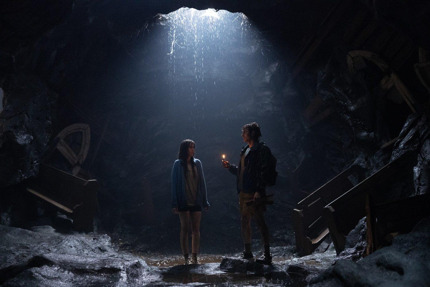 Zwei Personen stehen in einer dunklen Höhle, eine hält eine brennende Kerze. Licht fällt von einem Loch oben ein.