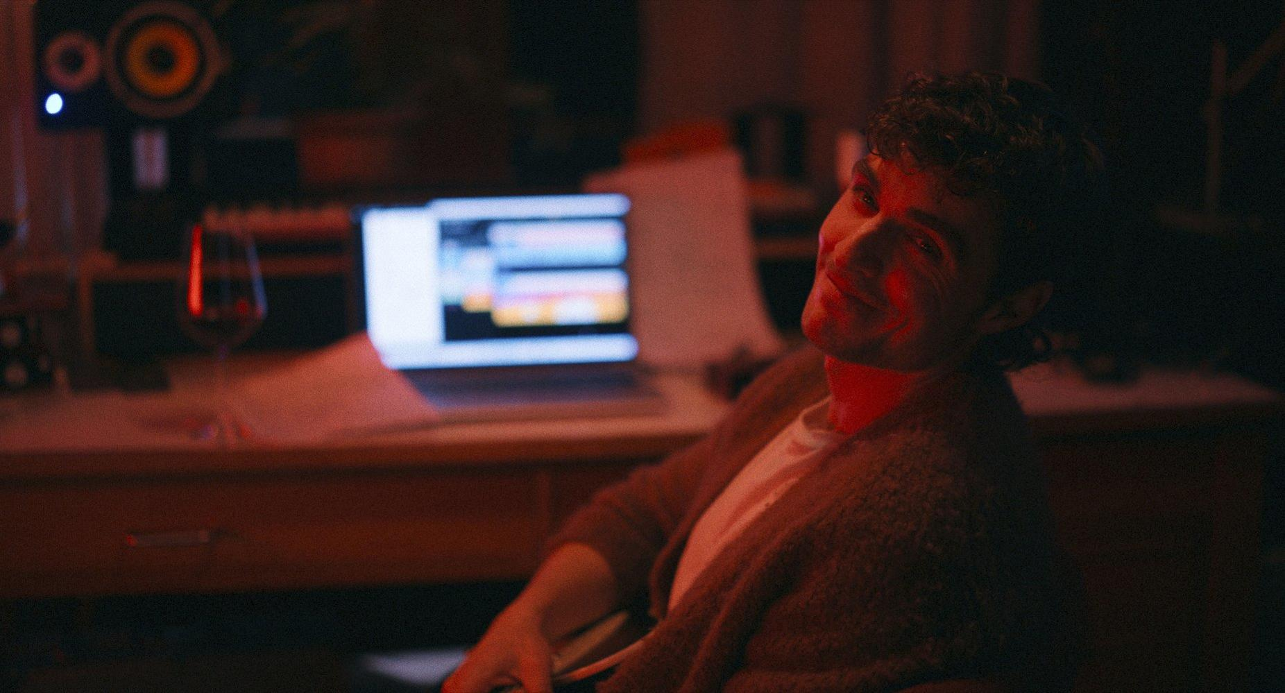 Ein Mann lächelt in einem schwach beleuchteten Studio. Laptop, Weinglas und Audioausrüstung sind zu sehen. Die Szene ist warm und einladend.