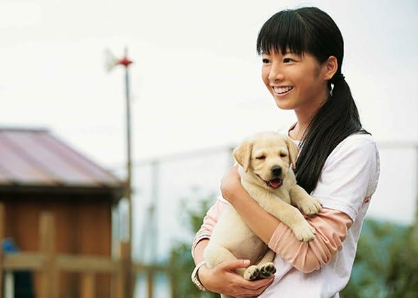 Eine lächelnde Frau hält einen gelben Labrador-Welpen. Die Frau hat dunkles Haar und trägt ein weißes Hemd. Der Welpe sieht zufrieden aus.