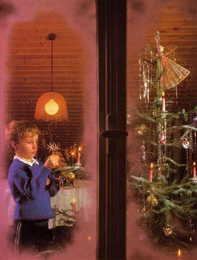 Ein Junge schmückt einen Weihnachtsbaum, der durch ein Fenster sichtbar ist. Drinnen sind Kerzen und Ornamente vorhanden. Draußen ist eine Lampe an.
