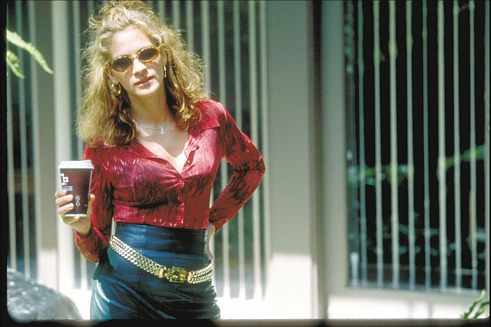 Eine Frau mit Sonnenbrille hält einen Kaffeebecher. Sie trägt ein rotes Hemd, einen schwarzen Rock und einen goldenen Gürtel. Der Hintergrund hat vertikale Linien.