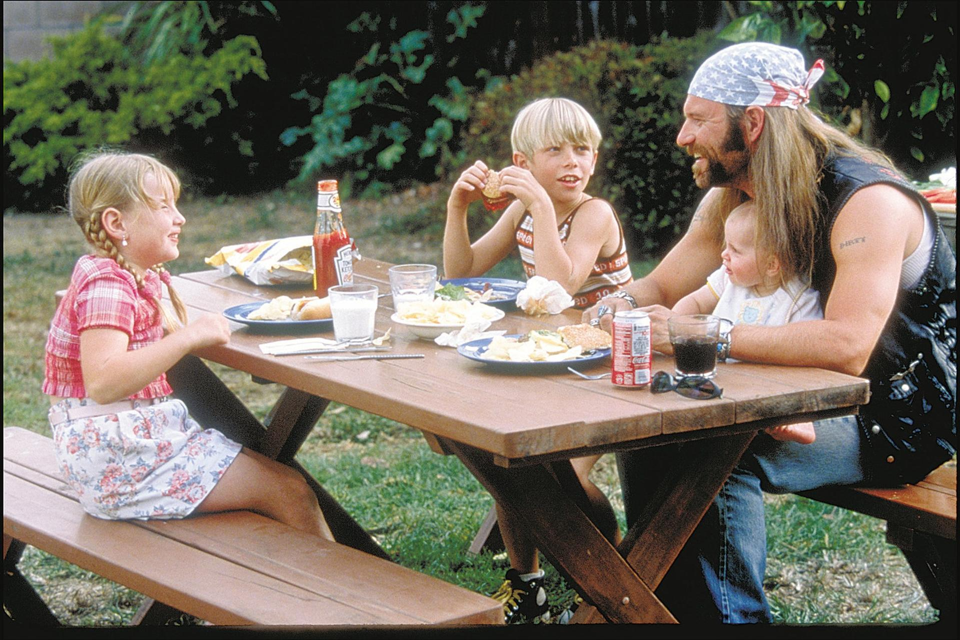 Familienpicknick. Kinder und Erwachsene an einem Picknicktisch essen Burger. Ein Mann mit Bandana hält ein Baby. Essen und Getränke auf dem Tisch.