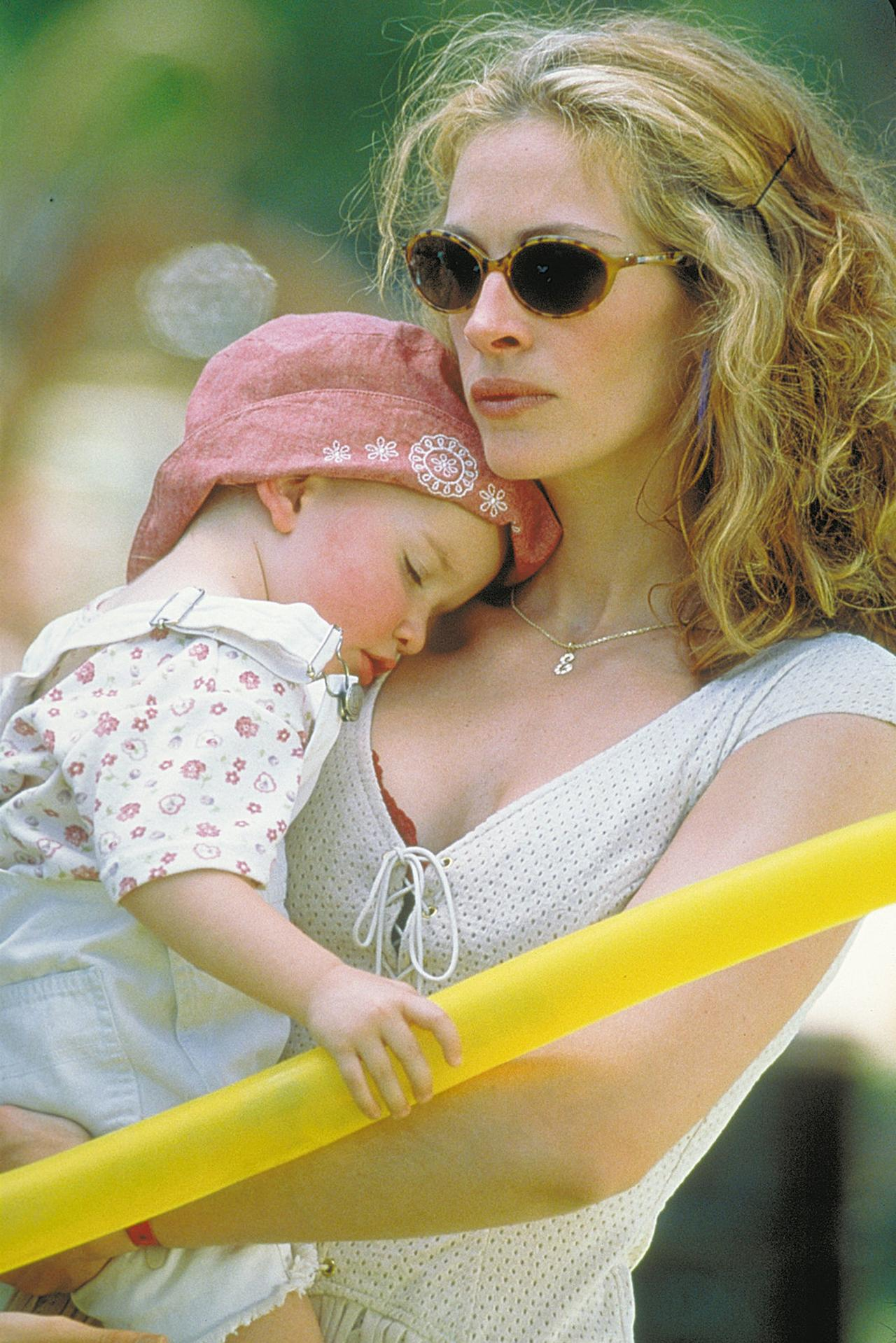 Frau mit Sonnenbrille hält schlafendes Kind mit rosa Hut. Gelbe Poolnudel im Vordergrund. Grüner Hintergrund.