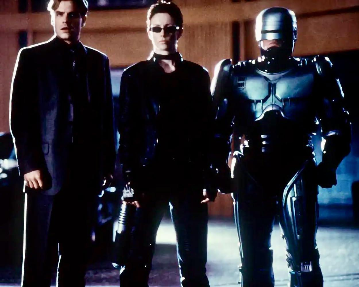 Drei Figuren stehen zusammen: ein Mann im Anzug, eine Frau in Schwarz und Robocop. Die Szene ist schwach beleuchtet.