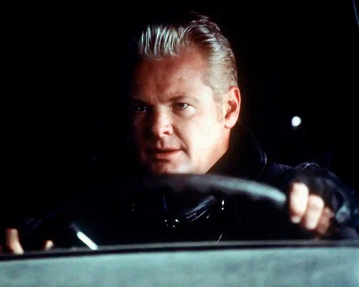 Ein Mann mit gegeltem, grauem Haar im Auto, hält das Lenkrad. Er trägt eine schwarze Lederjacke. Dunkler Hintergrund.