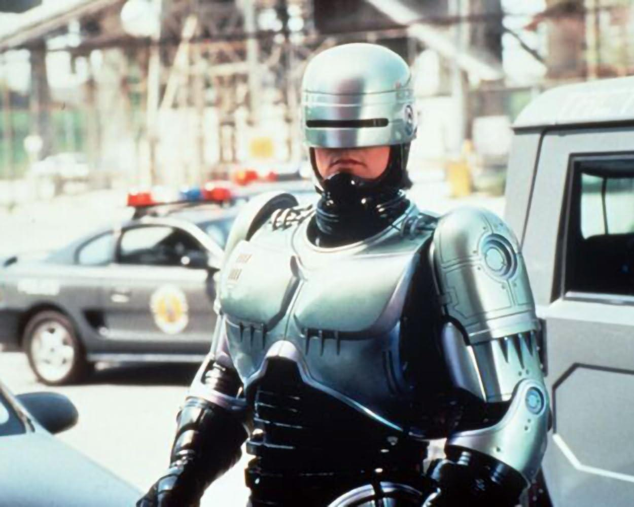 RoboCop steht vor einem Polizeiauto und trägt eine metallische Rüstung. Ein Polizeiauto mit Blinklicht ist hinter ihm.