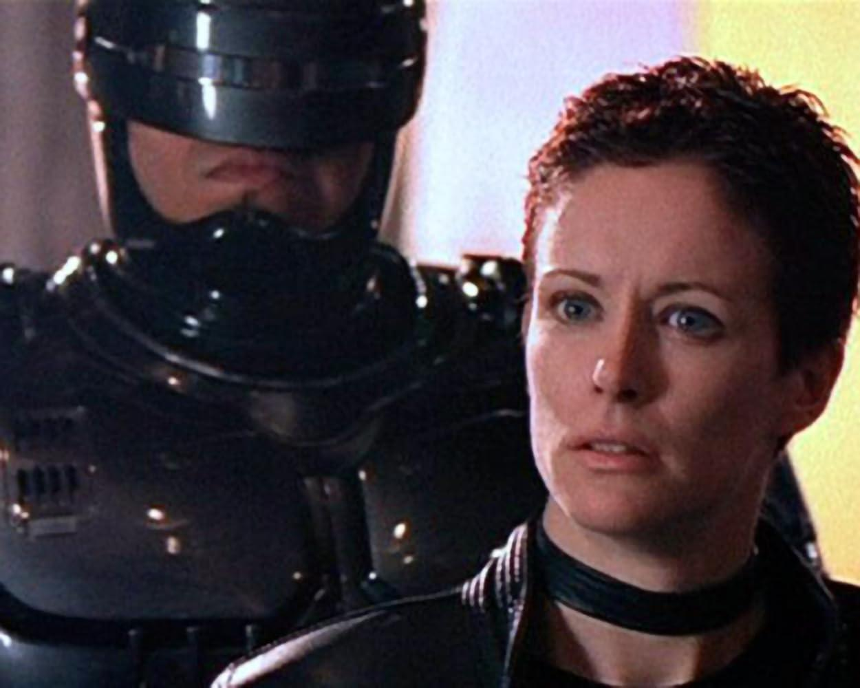 Eine Frau mit kurzen Haaren schaut in die Kamera. Hinter ihr ist ein Teil einer roboterartigen Figur in schwarzer Rüstung zu sehen.