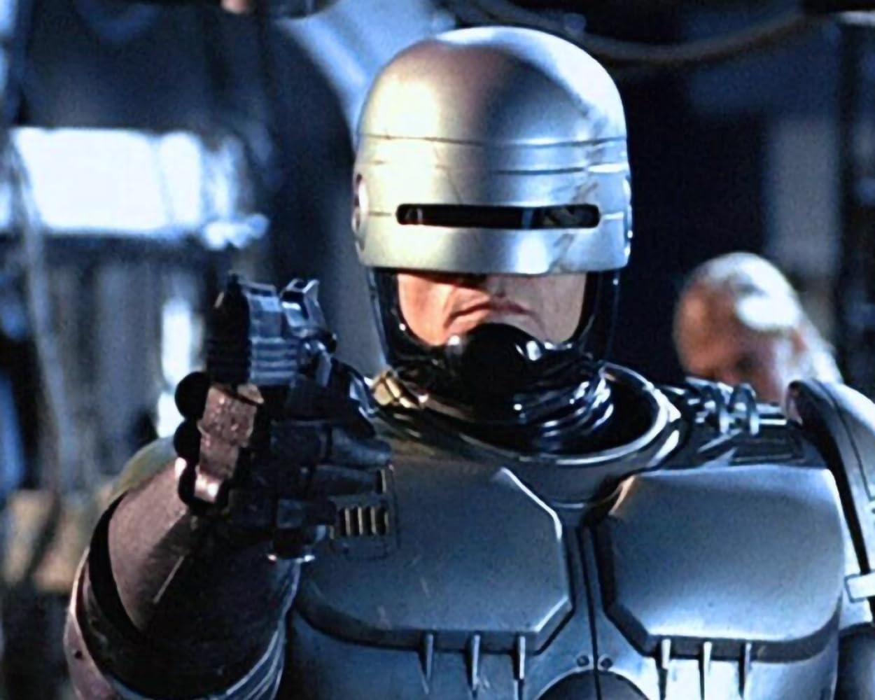 RoboCop in metallischer Rüstung, hält eine Waffe und zielt nach vorne. Im Hintergrund verschwommene Figuren und Metallstrukturen.