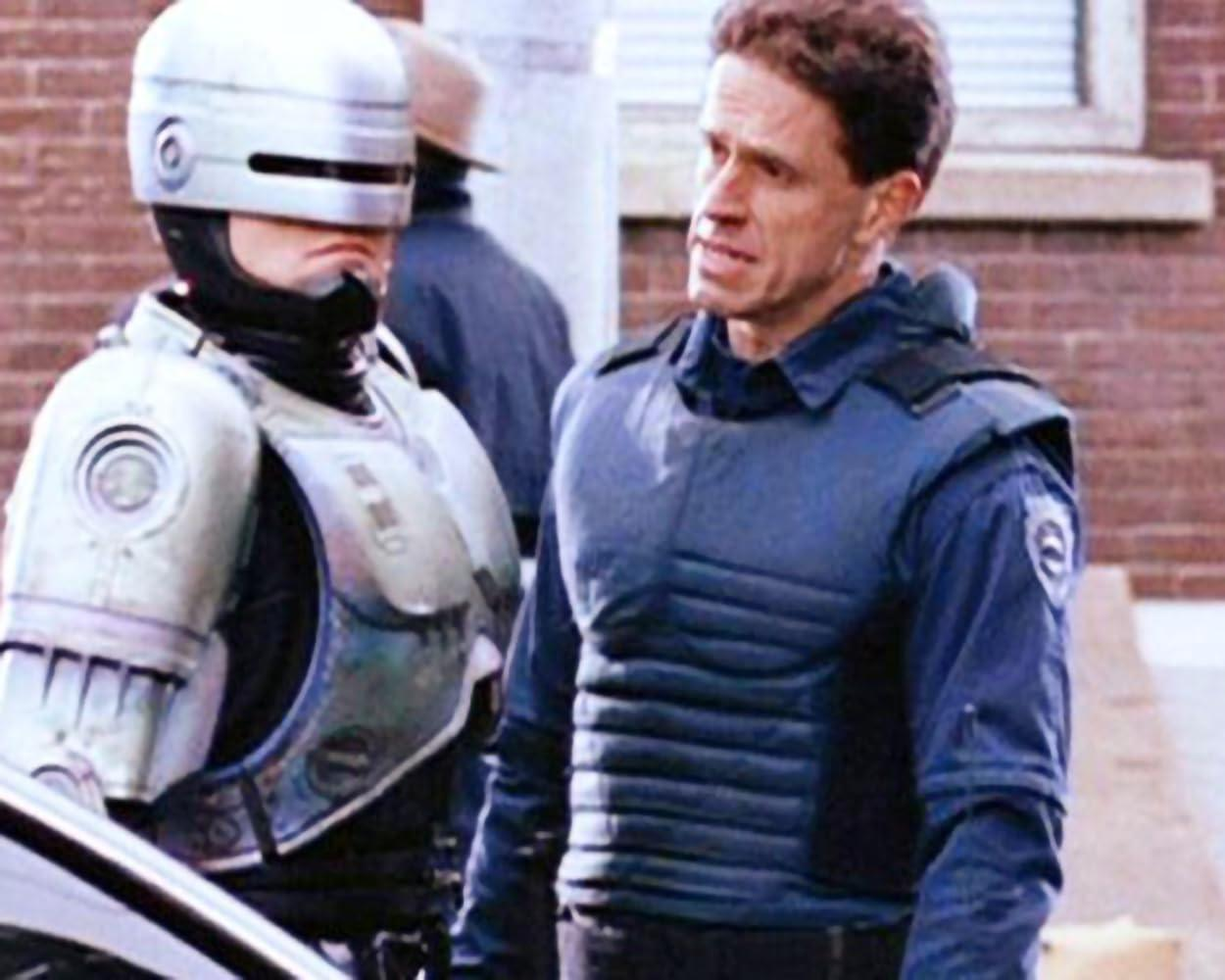 RoboCop und ein Offizier stehen zusammen. RoboCop ist metallisch. Der Offizier trägt blaue Uniform und eine kugelsichere Weste. Backsteinmauer im Hintergrund.