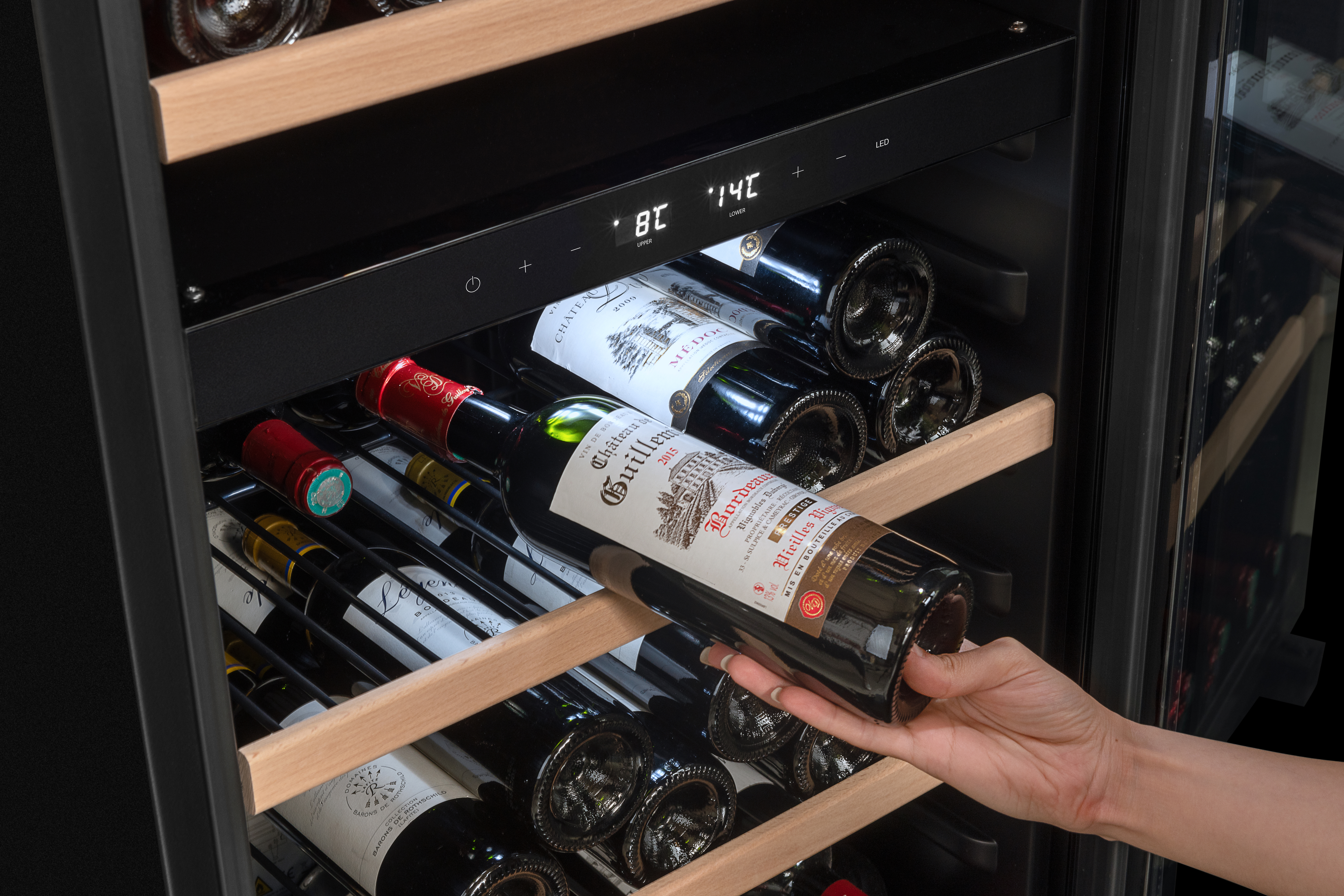 Eine Hand hält eine Flasche Rotwein in einem Weinkühler. Im Hintergrund sind weitere Weinflaschen zu sehen, mit Temperaturanzeige.