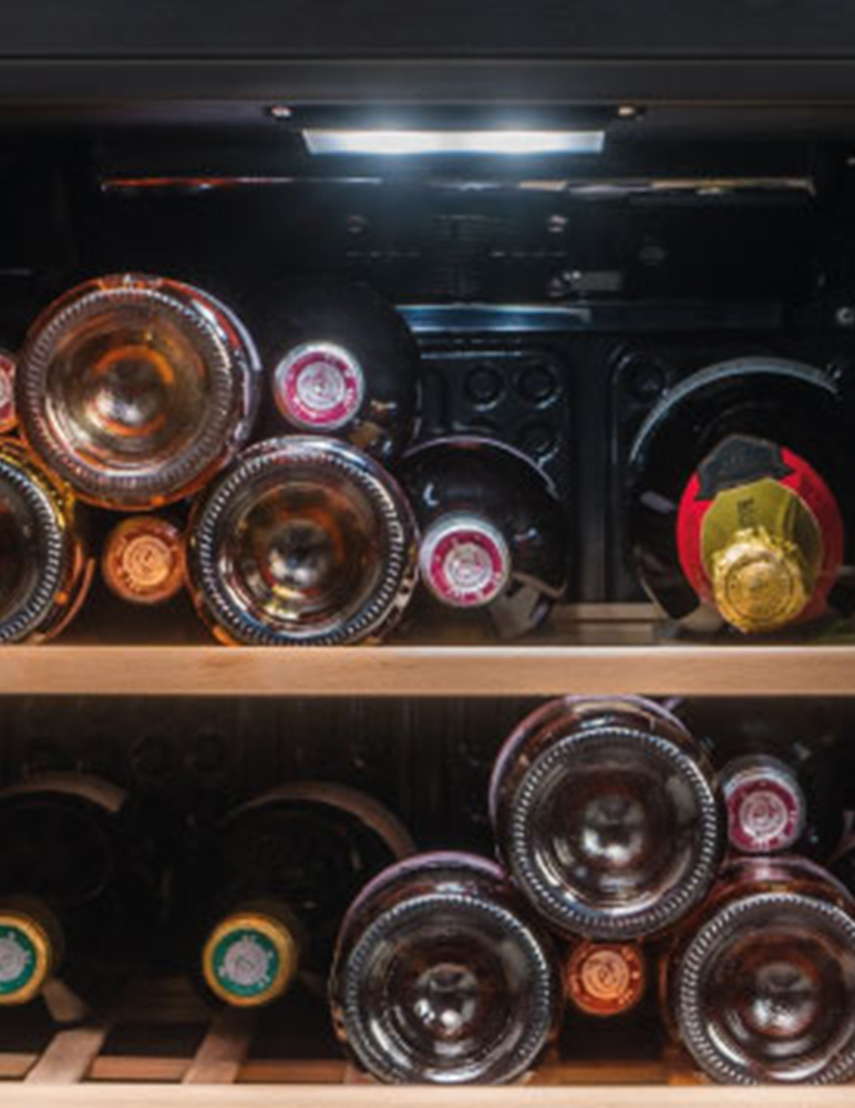 Ein Weinkühler voller Weinflaschen. Flaschen sind auf Regalen angeordnet. Einige haben rote Kappen, andere goldene. Der Hintergrund ist dunkel.